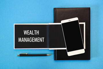A white notebook with black pages, a smartphone and a pen on a blue background. The inscription Wealth Management.