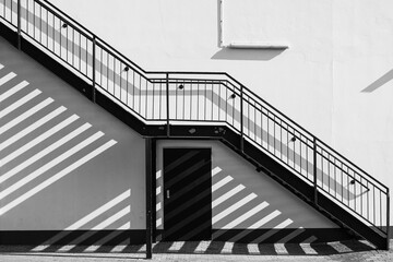 Eine Außentreppen zieht sch diagonal an der Hauswand entlang und wirft Schatten auf die Wand und den gepflasterten Boden - in schwarzweiß.