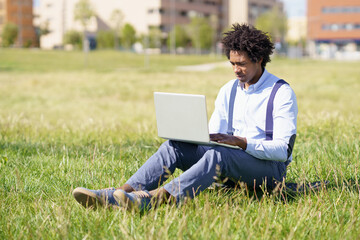 Black man with afro hair using his laptop sitting on skateboard on the grass of an urban park.