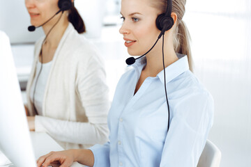 Blond woman call operator is using computer and headset for consulting clients online. Group of...