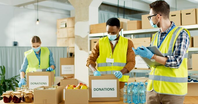 Portrait Of Young Caucasian Male Manager In Medical Mask Writing Checking Donation List. Mixed-race Volunteer Workers Working In Shipping Delivery Charitable Stock Organization Packing Donations Box