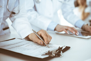 Unknown woman-doctors at work with patient at the background. Female physicians filling up medical documents or prescription while standing in hospital reception desk, close-up. Health care concept