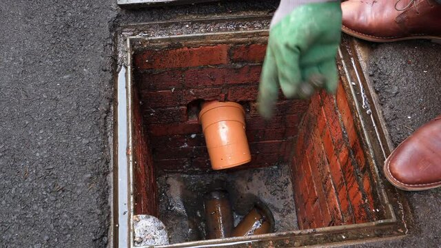 Inspecting drain sewer man with gloves on points to problem at open manhole
