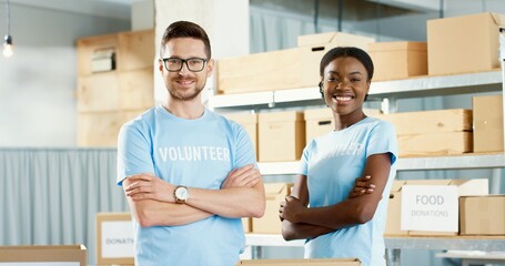 Portrait of happy cheerful young Caucasian handsome guy and beautiful African American woman volunteers standing in charity center stock looking at camera and smiling. Volunteering activity concept