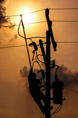 The silhouette of power lineman climbing on an electric pole with a transformer installed. And replacing the damaged hotline clamp, bail clamp, dropout and surge arrester that causes a power failure.