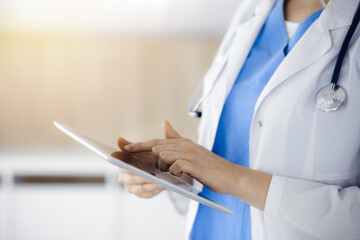 Unknown woman-doctor standing in sunny clinic and using tablet pc, close-up. Data and best service in medicine