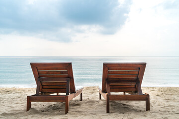 Fototapeta premium Back view of two wooden sunbeds together on the beautiful beach in the morning, the background of a wide-open view of sea and horizon, blank space for copy.