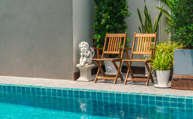 Two vintage wooden chairs together beside swimming pool with small garden decoration by many tree in pots, daylight image.