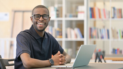 Young African Man with Laptop Smiling at Camera in Library
