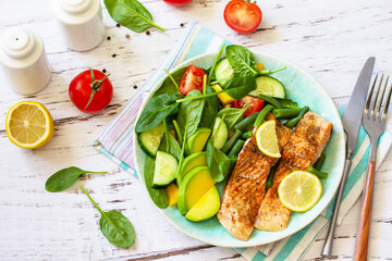 Grilled salmon fillet with spinach, cucumber, tomato and avocado salad on a white wooden table.