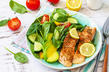 Grilled salmon fillet close-up with spinach, cucumber, tomato and avocado salad on a white wooden table.