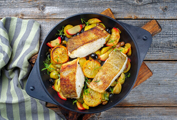 Modern Style traditionelles gebratenes Skrei Kabeljau Fisch Filet mit Bratkartoffel, Früchten und Oliven serviert als Draufsicht in einer gußeisernen Design Pfanne