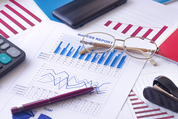 Business graphs, charts, data tables, business report, pen, glasses, calculator, smartphone.