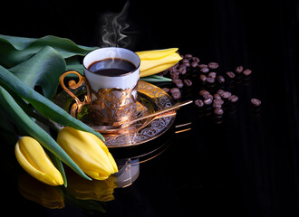 Steaming golden coffee cup, yellow tulip flowers and coffee beans on black background with reflection