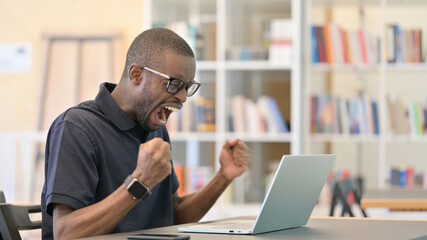 Fototapeta na wymiar African Man Celebrating Success on Laptop in Library