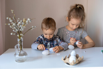 Obraz premium Brother and sister play at the table. A boy and a girl are preparing for Easter. Children's educational activities at home. The joint work of a brother and sister. Coloring decorative eggs.