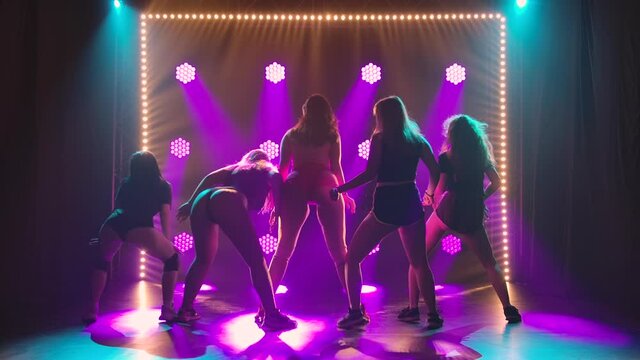 Rear view of a group of seductive dancers shaking their butts in short shorts. Silhouettes of young women twerking their asses moving their hips in a dark studio with colored lights. Slow motion.