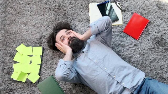 Young bearded man lying on the floor, surrounded by computer and documents, the man is very tired and exhausted, laptop is full of reminder notes.