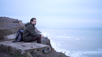 Sad lonely young man, with backpack, sits on a cement block at the edge of cliff above storm sea,...