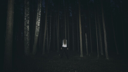 Scary person with long hair and bloody white tshirt standing in a dark forest. Zombie man walking in misty night forest.