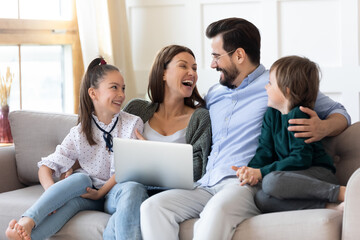 Overjoyed young parents have fun with two small children use modern computer gadget together. Happy Caucasian family laugh talk speak on video call on laptop. Technology, virtual event concept.