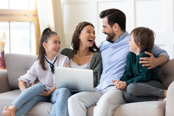 Fototapeta premium Overjoyed young parents have fun with two small children use modern computer gadget together. Happy Caucasian family laugh talk speak on video call on laptop. Technology, virtual event concept.