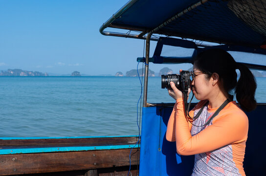 young asian woman weraing swimsuit using camera to take photo of island and blue sea while travel on long tail boat on summer vacation