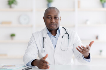 Patient's point of view of medical consultation. Black male doctor gesturing to camera in his office