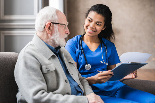 Happy young skilled female general practitioner in blue medic uniform visiting disabled smiling middle aged senior patient, showing health analysis on paper, satisfied with treatment results.