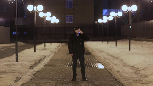 Handsome man in a black coat talking on a smartphone at night on the street in winter.