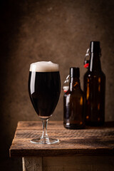 Glass of dark beer with foam head on dark wooden background with empty bottles and beer snacks