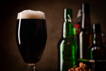 Glass of dark beer with foam head on dark wooden background with empty bottles and beer snacks