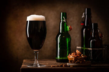 Glass of dark beer with foam head on dark wooden background with empty bottles and beer snacks