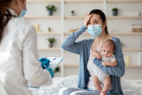 Mother In Protective Mask Holding Newborn Baby, Explaining Illness Symptoms To Doctor