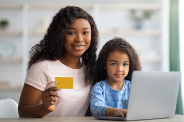 Online shopping. Happy black woman and her daughter using credit card and laptop to buy goods on web from home