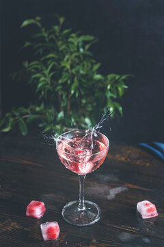 Splash of pink rose champagne in Elegant glass with frozen raspberries in ice cube on dark wooden table surface