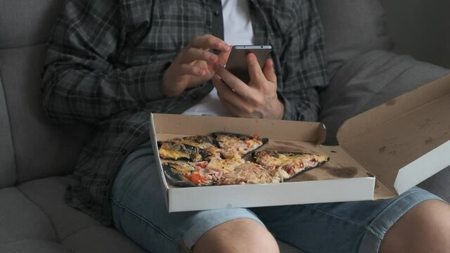Man is taking photos of pizza in box on his knees using smartphone, closeup view. Unusual pizza with black dough. He is sitting on sofa at home, food blogger, social media life concept.