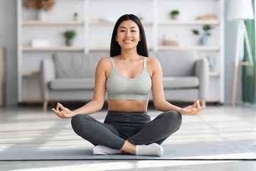 Fototapeten Domestic Yoga. Happy young asian woman meditating at home in lotus position © Prostock-studio