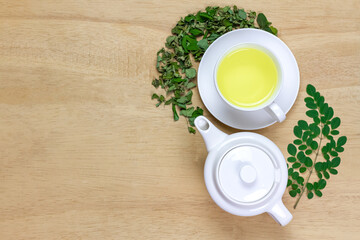 Moringa Tea in white cup and fresh green leaf with teapot on wooden background, top view. Moringa oleifera tropical herb healthy lifestyle concept.