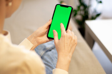 Woman at home, on the couch, using a smartphone with a green mockup screen. Girl using a mobile phone on the Internet. Point of view shot