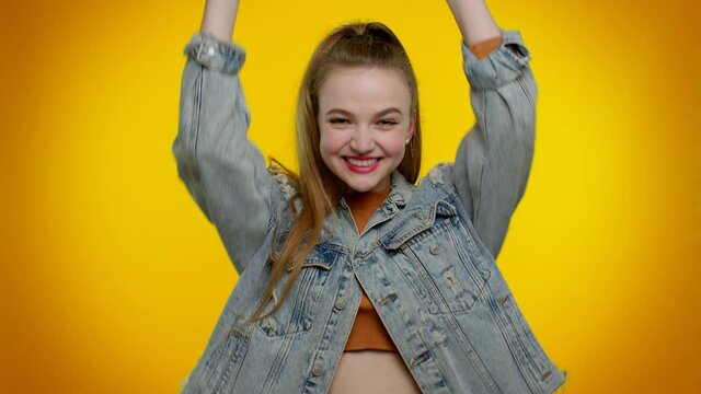 Pretty stylish teen girl in denim jacket shouting, raising fists in gesture I did it, celebrating success, winning and goal achievemen. Young adult woman indoor on yellow studio wall background