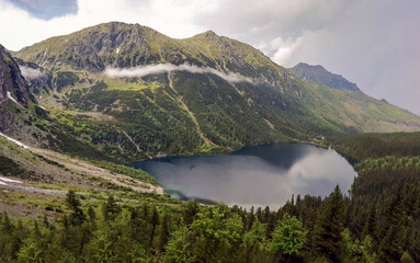 Fototapeta na wymiar Wide angle top view of Morskie Oko naturally formed lake pond in Tatra Mountains in Poland. High mountain landscape with dramatic clouds covered with snow and trees at national park High Tatras Europe