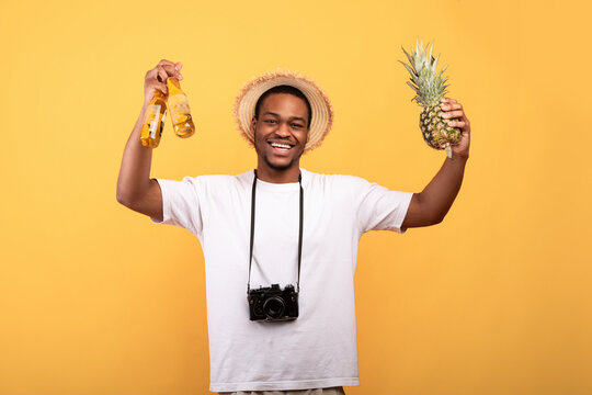 Portrait of handsome black guy in summer clothes with camera holding beer bottles and pineapple, looking at camera