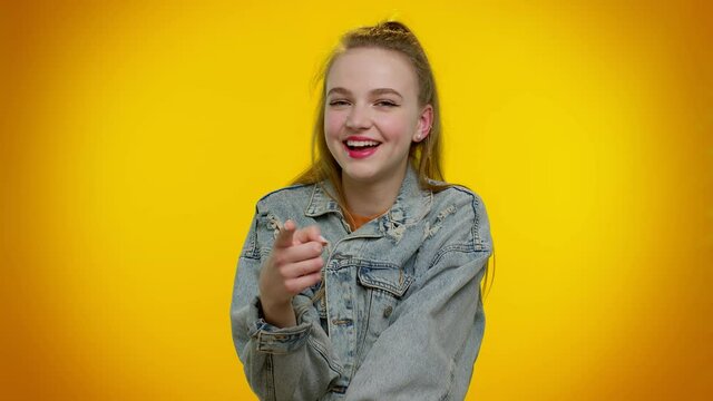 Amused teen stylish girl in denim jacket pointing finger to camera, laughing out loud, taunting making fun of ridiculous appearance, funny joke. Young woman posing on yellow studio wall background