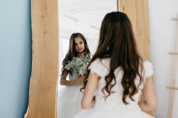 preteen flower girl in white dress with a bouquet of white gypsophila stands near the mirror and...