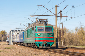 A powerful green two-section locomotive pulls blue passenger cars. Sunny spring weather. Close-up view.