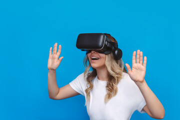 A young blonde delighted astounded woman in a white t-shirt looks into black virtual reality glasses on blue background. VR gadget concept. Modern technology, bright colorful world. Positive emotions