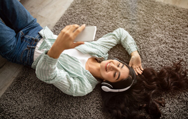 Happy Indian lady in headphones lying on floor and listening to music on mobile device at home,...