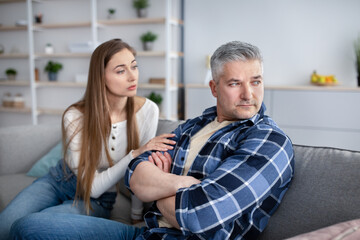 Family conflict concept. Upset mature man sitting on couch, his wife trying to make peace after fight at home