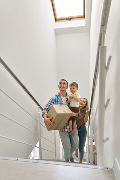 Happy family couple new home owners with toddler kid child son carrying cardboard boxes going up stairs on moving day arriving into new home. Relocation, real estate property purchase, removal.